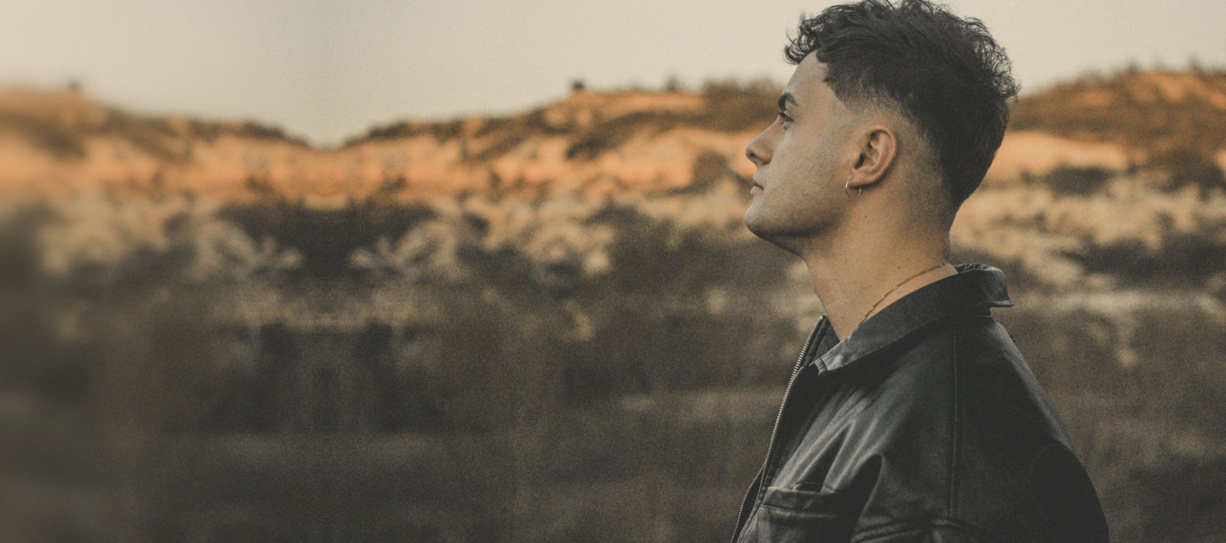 Man in a black leather jacket in profile, looking across red-earth hills