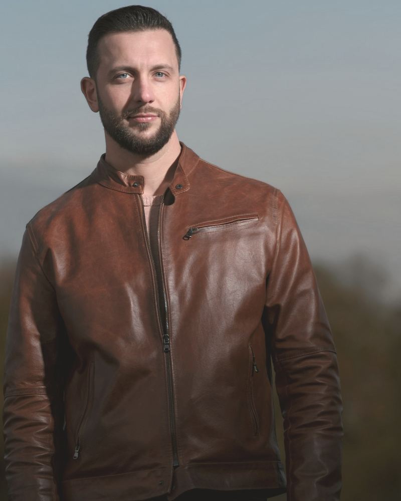 A man in a cognac leather racer jacket looks into the camera against a dusk sky