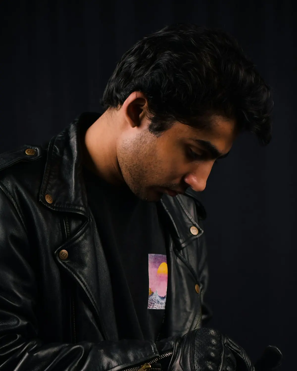 A black leather biker jacket with metal snaps, worn by a man looking down, in a dark studio